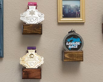 May include: A display of medals and framed photographs on a beige wall. The medals are presented on small wooden blocks. A blue framed photo is on the left, and a gold framed photo is in the centre.