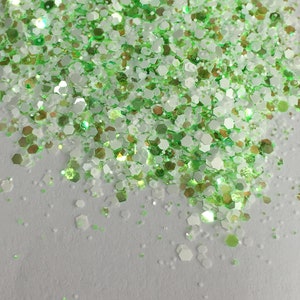 May include: Close-up of a pile of glitter in shades of green, white, and gold. The glitter particles are hexagonal and of varying sizes, creating a textured appearance. The background is a plain white surface.
