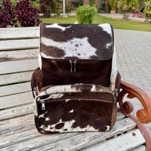 Puede incluir: Una mochila de piel de vaca con cierre de solapa y bolsillo delantero con cremallera. La bolsa presenta un estampado moteado marrón y blanco, con ribetes marrones y cremalleras plateadas. La mochila está sobre un banco de madera.