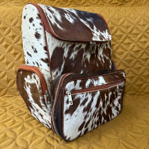 May include: A brown and white cowhide leather backpack with orange zipper accents. The backpack has two large compartments and a smaller front pocket.
