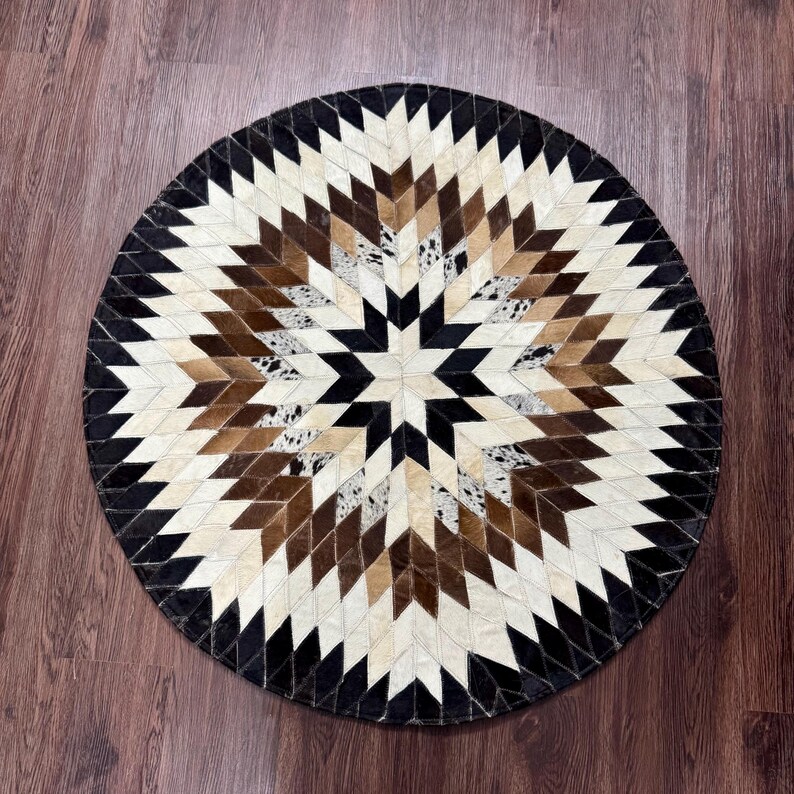 Pode incluir: Tapete redondo com um design geom&eacute;trico de estrela. O tapete apresenta um padr&atilde;o de losangos em tons de preto, castanho, creme e branco. O tapete &eacute; feito de um material natural, provavelmente pele ou couro.