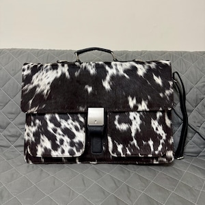 May include: A black and white cowhide leather briefcase with a top handle and a shoulder strap. The bag has a flap closure with a silver-tone buckle and two front pockets. The briefcase is designed for carrying documents and other essentials.