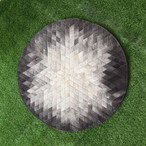 Könnte beinhalten: Ein runder Teppich mit einem geometrischen Muster aus kleinen rautenförmigen Fliesen in verschiedenen Grau-, Anthrazit- und Cremetönen. Das Design strahlt von der Mitte aus und erzeugt einen sternartigen Effekt mit einem Farbverlauf.