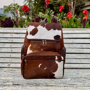 May include: A brown and white cowhide backpack with a flap closure and multiple zippered compartments. The bag has a top handle and adjustable straps. The backpack is sitting on a wooden bench outdoors.