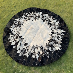 Peut inclure: Un tapis rond avec un motif géométrique. Le tapis présente une section centrale blanche entourée de sections à motifs noirs et blancs, et une bordure noire. Le tapis est fait d'une matière naturelle et est posé sur une surface herbeuse verte.
