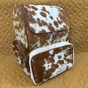 Puede incluir: Una mochila de piel de vaca marrón y blanca con cierre de solapa y bolsillo delantero. La mochila tiene ribetes marrones y cremalleras plateadas. La bolsa está sobre una superficie beige.