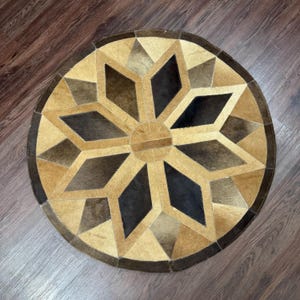 Puede incluir: Alfombra redonda con un diseño geométrico de estrella. La alfombra presenta un círculo central y un patrón de estrella en tonos marrones, beige y negro. Está hecha de un material natural, probablemente piel o cuero, y tiene un borde marrón oscuro.