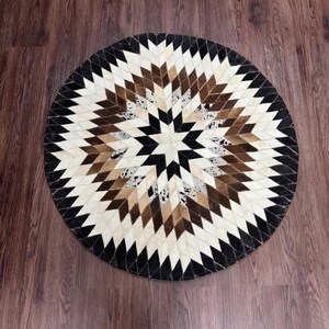 Pode incluir: Um tapete redondo com um padr&atilde;o geom&eacute;trico. O tapete apresenta um design central em forma de estrela em tons de castanho, preto e creme. A borda externa &eacute; preta e o tapete parece ser feito de couro ou material semelhante.
