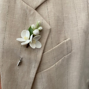 Puede incluir: Un prendedor floral blanco con hojas y capullos verdes, sujeto a una blazer beige con finas rayas. El prendedor presenta dos flores abiertas y un alfiler plateado. La blazer tiene solapas con muescas y un bolsillo.