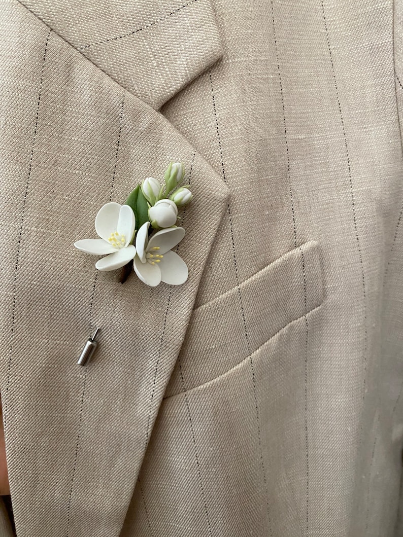 Puede incluir: Un prendedor floral blanco con hojas y capullos verdes, sujeto a una blazer beige con finas rayas. El prendedor presenta dos flores abiertas y un alfiler plateado. La blazer tiene solapas con muescas y un bolsillo.
