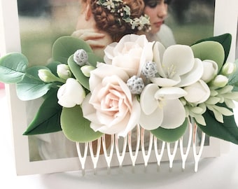 May include: A white and blush floral hair comb with a silver comb. The comb is decorated with white roses, white flowers, and green leaves.