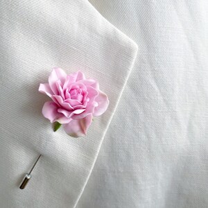 May include: A pink fabric rose flower with a silver pin attached to the back. The rose is pinned to a white fabric surface.