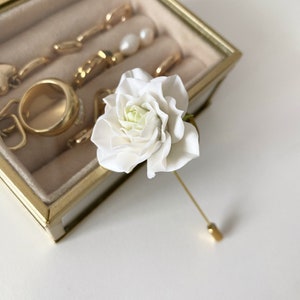May include: A white flower pin with a gold stem and pin. The flower is a single bloom with a white center and white petals.
