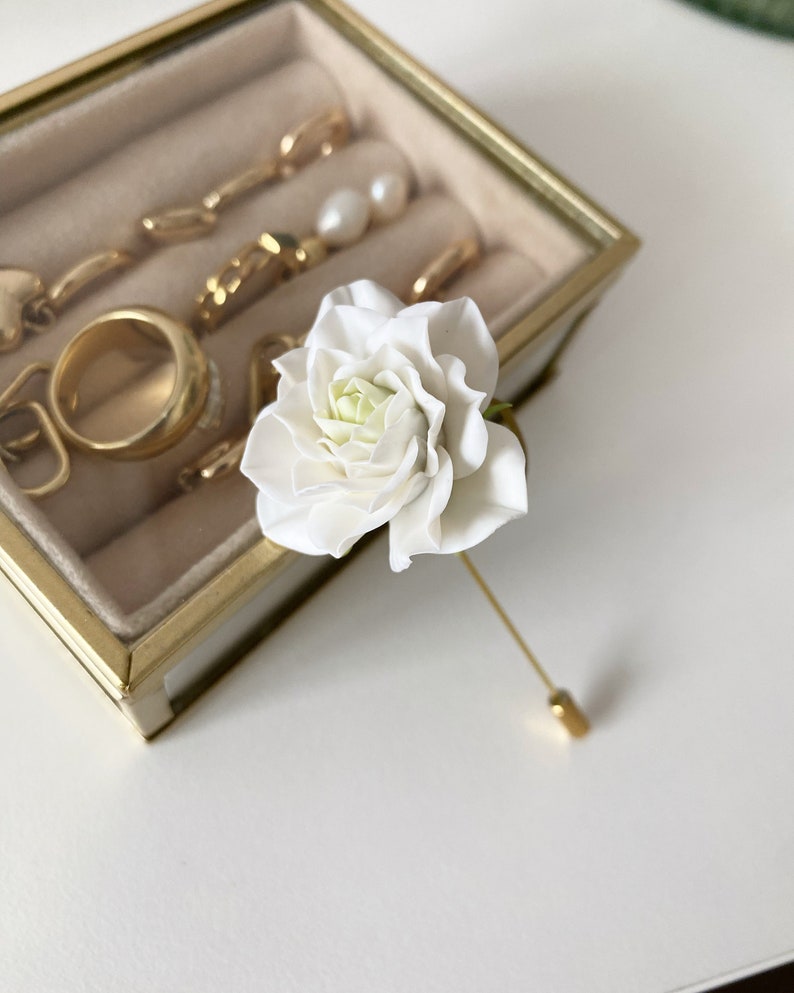 May include: A white flower pin with a gold stem and pin. The flower is a single bloom with a white center and white petals.