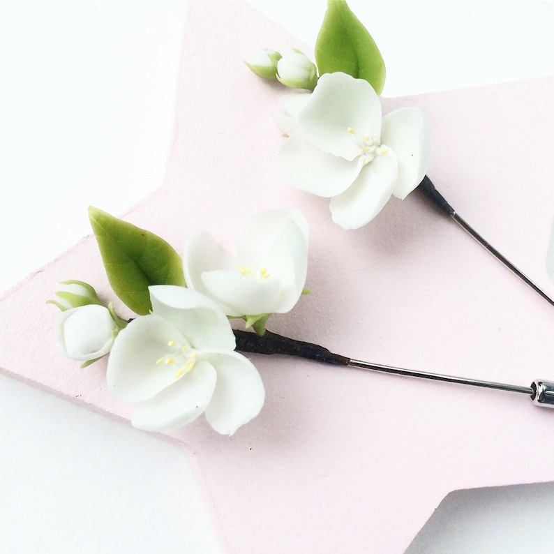 Puede incluir: Dos prendedores florales con flores blancas, centros amarillos y hojas verdes. Los prendedores tienen un tallo negro y un respaldo de alfiler plateado. Se colocan sobre un fondo rosa claro en forma de estrella.