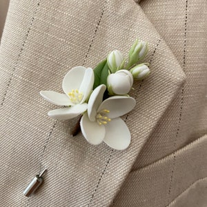 Puede incluir: Un prendedor floral blanco con hojas verdes y un tallo marr&oacute;n, sujeto a una blazer beige con rayas finas. El prendedor presenta flores blancas detalladas con centros amarillos y capullos sin abrir. Un alfiler plateado asegura el accesorio floral.