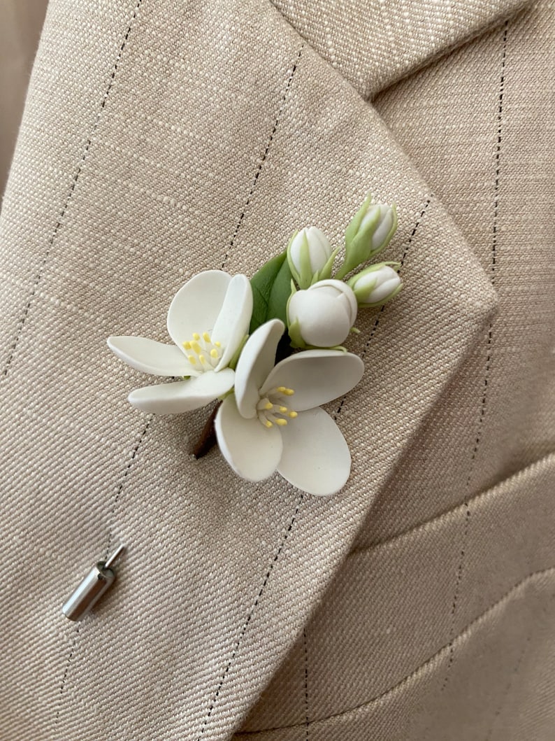 Puede incluir: Un prendedor floral blanco con hojas verdes y un tallo marr&oacute;n, sujeto a una blazer beige con rayas finas. El prendedor presenta flores blancas detalladas con centros amarillos y capullos sin abrir. Un alfiler plateado asegura el accesorio floral.