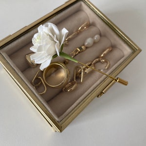 May include: A gold jewelry box with a white flower and gold jewelry inside. The box has a clear glass lid and a gold metal frame.