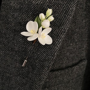 Puede incluir: Un prendedor de solapa de flores blancas con un respaldo de alfiler plateado. El prendedor presenta dos flores abiertas, capullos y hojas verdes. El prendedor est&aacute; sujeto a una blazer de espiga gris oscuro.