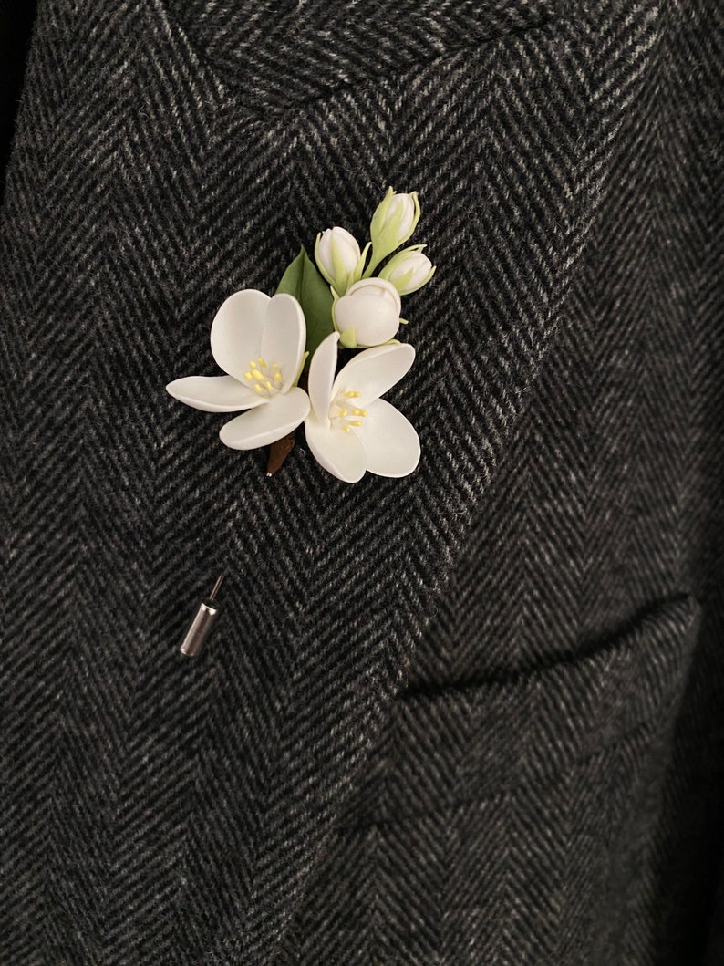 Puede incluir: Un prendedor de solapa de flores blancas con un respaldo de alfiler plateado. El prendedor presenta dos flores abiertas, capullos y hojas verdes. El prendedor est&aacute; sujeto a una blazer de espiga gris oscuro.
