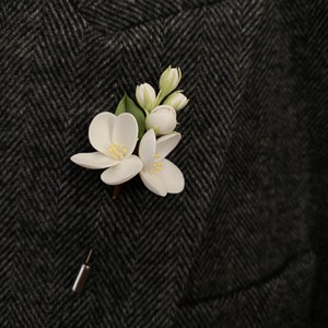 Puede incluir: Un prendedor floral blanco con hojas verdes y un respaldo de alfiler plateado. El prendedor est&aacute; sujeto a una tela oscura con estampado de espiga, probablemente una chaqueta de traje. Las flores tienen centros amarillos.