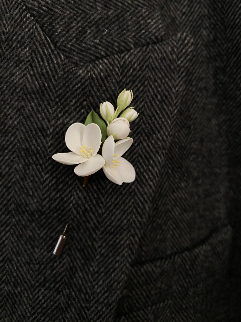 Puede incluir: Un prendedor floral blanco con hojas verdes y un respaldo de alfiler plateado. El prendedor est&aacute; sujeto a una tela oscura con estampado de espiga, probablemente una chaqueta de traje. Las flores tienen centros amarillos.