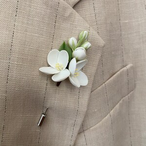 Puede incluir: Un prendedor floral blanco con hojas verdes y detalles amarillos, sujeto a una blazer beige con finas rayas. El prendedor presenta un grupo de flores y capullos delicados, a&ntilde;adiendo un toque de elegancia a la prenda.