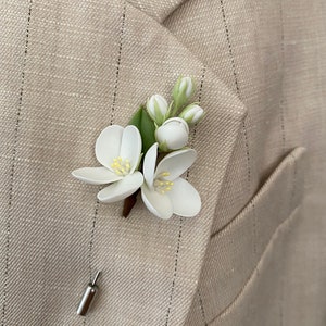 Puede incluir: Un prendedor floral blanco con hojas y capullos verdes, sujeto a una chaqueta de traje beige con rayas finas. El prendedor presenta dos flores abiertas con centros amarillos y un respaldo plateado.