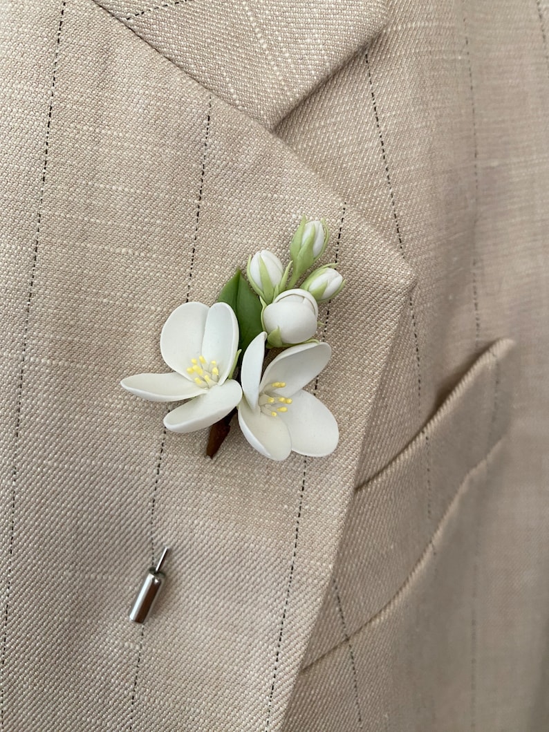 Puede incluir: Un prendedor floral blanco con hojas y capullos verdes, sujeto a una chaqueta de traje beige con rayas finas. El prendedor presenta dos flores abiertas con centros amarillos y un respaldo plateado.