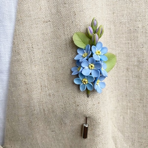 May include: A blue and green forget-me-not flower boutonniere with a silver pin.