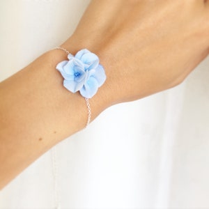May include: A delicate silver chain bracelet with a light blue flower charm. The flower is made of polymer clay and has a realistic appearance.