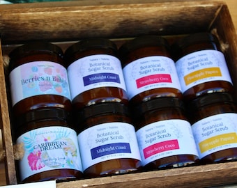 May include: Eight amber glass jars of Botanical Sugar Scrub arranged in a wooden crate. Labels feature various scents including Berries n Bubbly, Midnight Coast, Strawberry Coco, Pineapple Papaya, and Caribbean Dream, each marked with a net weight of 8 oz.
