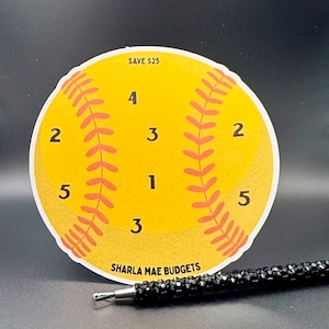 May include: A yellow softball-shaped budget tracker with red stitching and numbered sections. The text "SAVE $25" is at the top, and "SHARLA MAE BUDGETS" is at the bottom. A black pen with crystal accents rests nearby.