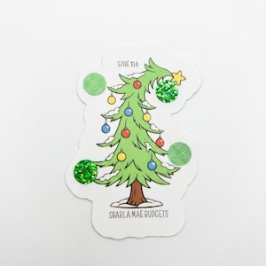 May include: A die-cut sticker featuring a cartoon Christmas tree with a brown trunk, green branches, and snow accents. The tree is decorated with colorful ornaments and two green glitter circles. Text reads SAVE $14 and SHARLA MAE BUDGETS.