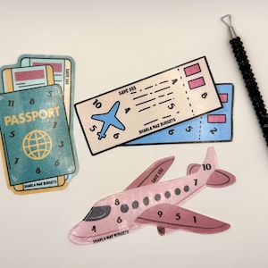 May include: A collection of travel-themed stickers and a black beaded tool. The stickers include a passport, airplane, and tickets, all with the text "SHARLA MAE BUDGETS" and numbers. The airplane is pink and the tickets are blue.