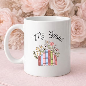 May include: White ceramic mug with the text "Ms. Silvia" in black script. The mug features a colorful illustration of books and flowers. The mug is on a pink surface with a floral background.
