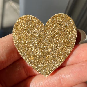 Puede incluir: Decoración en forma de corazón con purpurina dorada. El corazón está cubierto de purpurina dorada brillante, que capta la luz. La forma es lisa y simétrica, ideal para manualidades o adornos. El corazón se sostiene en una mano.