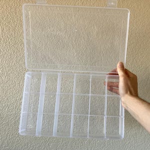 May include: A clear plastic storage container featuring a hinged lid and a rectangular base divided into fifteen individual compartments by fixed vertical and horizontal dividers, designed for organizing small items and craft supplies.
