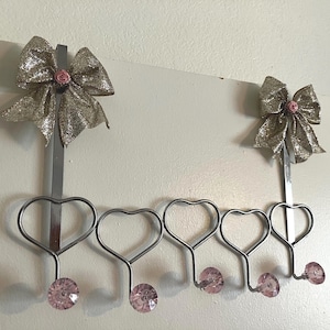 May include: A decorative over-the-door hanger with five heart-shaped hooks. The hanger is silver-toned metal and features a gold glitter bow with a pink rose in the center. Each hook has a pink crystal-like accent.