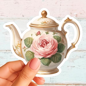May include: A die-cut sticker featuring a cream-colored teapot with a large pink rose illustration, green leaves, and gold trim accents on the handle, spout, lid, and base. The teapot has a rounded body and a matching lid with a gold knob.