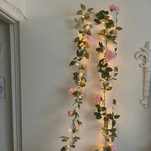 Puede incluir: Dos guirnaldas de rosas artificiales con pequeñas luces LED blancas cálidas. Las rosas rosas y las hojas verdes están colgadas verticalmente contra una pared blanca, creando un ambiente romántico y decorativo. Las luces están espaciadas uniformemente.