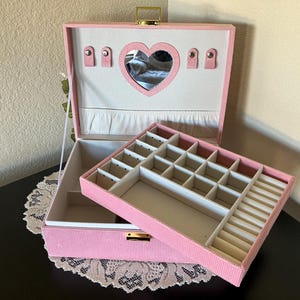 May include: Pink jewelry box with a heart-shaped mirror and multiple compartments. The box is open, revealing a removable tray with various storage slots. The exterior is a textured pink fabric, and the interior is lined with a lighter pink material.