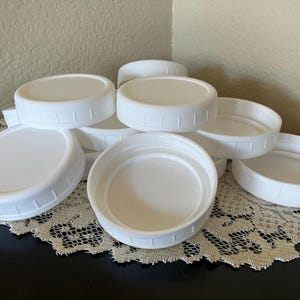 以下が含まれることがあります： 滑らかな上面とリブ付きの縁を持つ、白いプラスチック製の瓶の蓋のコレクション。これらの円形の蓋は、しっかりと閉まるように内側にネジ山が施されており、上面と底面の両方が見えるように積み重ねて配置されています。