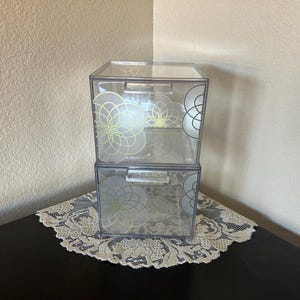 May include: Two clear, stackable storage boxes with decorative floral designs. Each box has a rectangular shape with a hinged lid and a silver handle. The floral design is in a light gold color.