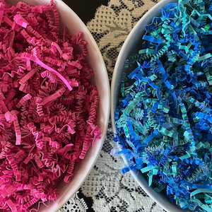 Pode incluir: Duas tigelas brancas cheias de papel crepom colorido. Uma tigela é preenchida com papel rosa brilhante, e a outra com papel azul e verde-azulado. O papel é desfiado e tem uma aparência texturizada. Ideal para embalagens de presente ou artesanato.