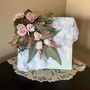 May include: A decorative box with a light blue painted surface, decorated with white lace and a floral arrangement of pink roses, green leaves, and sprigs. The box sits on a lace doily.