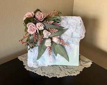 Puede incluir: Una caja decorativa con una superficie pintada de color azul claro, adornada con encaje blanco y un arreglo floral de rosas rosas, hojas verdes y ramitas. La caja se encuentra sobre un tapete de encaje.