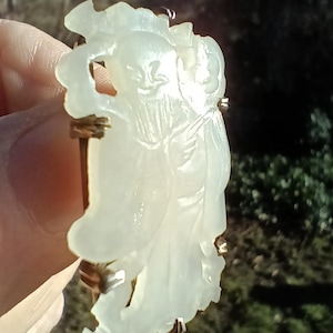 May include: A light green jade carving of a figure with flowing robes, set in a gold-toned metal frame. The carving is held in a hand, with a blurred outdoor background. The carving has intricate details and a smooth surface.