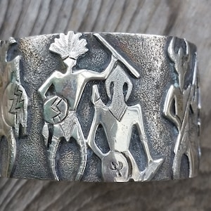 May include: A silver bracelet with a detailed design of stylized figures and geometric patterns. The bracelet is made of a textured metal and has a slightly worn look.