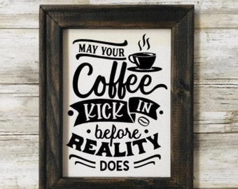 May include: A framed sign with the text "May your coffee kick in before reality does" in black lettering on a white background. The sign features a stylized coffee cup with steam rising from it.